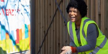 A woman in a bright green vest and gloves smiling at someone off camera as she works to clean up her neighborhood.