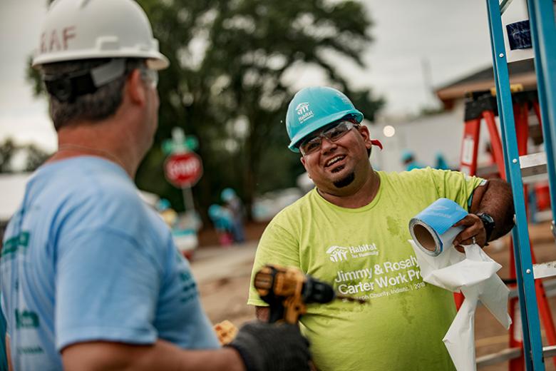 Man in green shirt and hard hat smiling while working on build site.